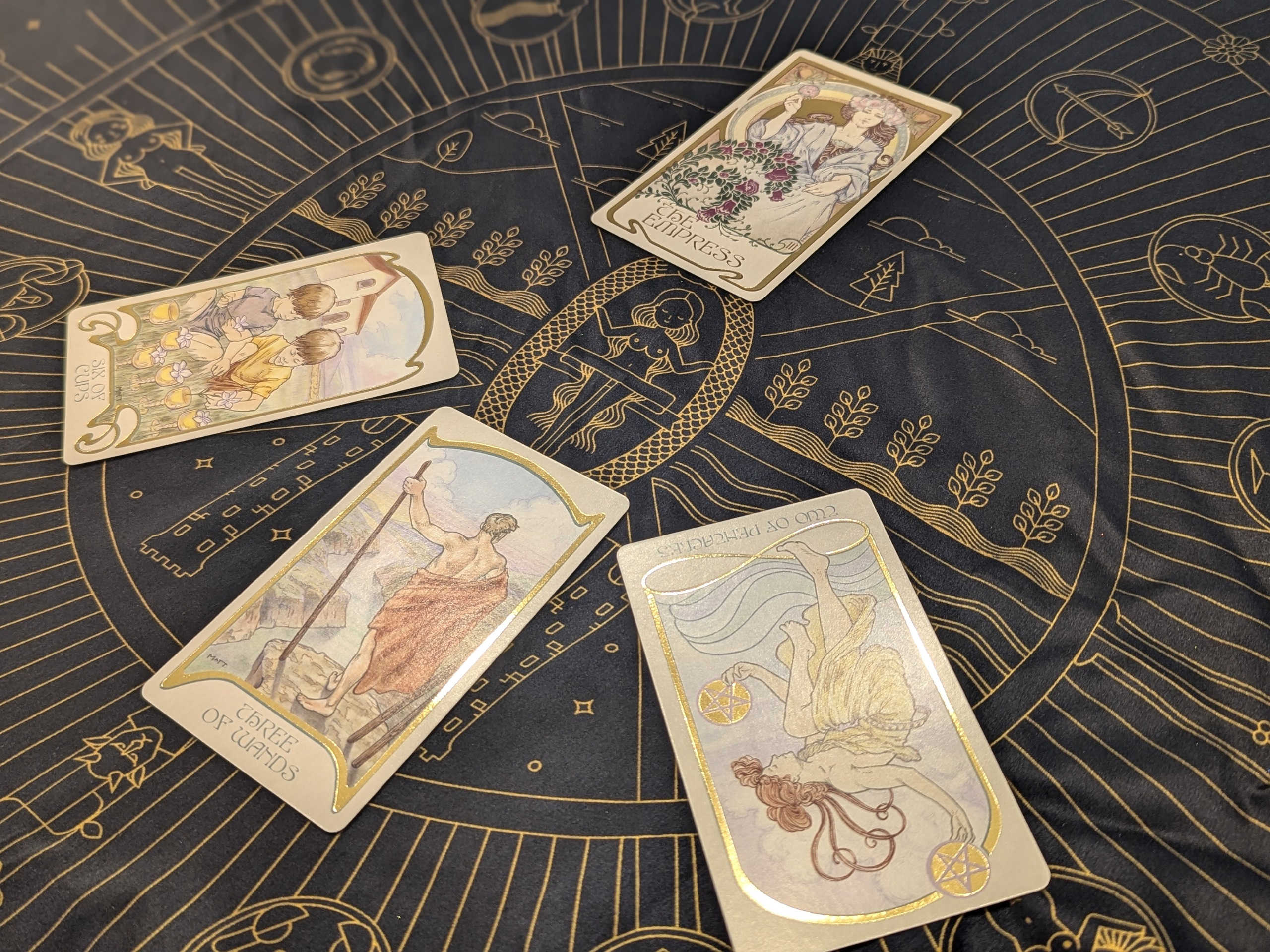 A spread of tarot cards on top of a reading cloth. There are four cards. The empress sits atop a spread of three cards below it: The six of cups, three of wands and two of pentacles reversed. The cloth features images of tarot cards and the zodiac.