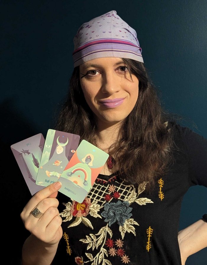 A picture of Luna. She is dressed in a black shirt with floral embriodery and a purple veil. She is smiling with purple lipstick. In her right hand, she holds three cards: the Fool reversed, the High Priestess, and Temperance.